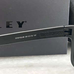 May include: Close-up of black Oakley Holbrook sunglasses. The frame features the model number OO9102-90 55 18 137. The word "HOLBROOK" is printed on the arm. The background is a black box with the Oakley logo.