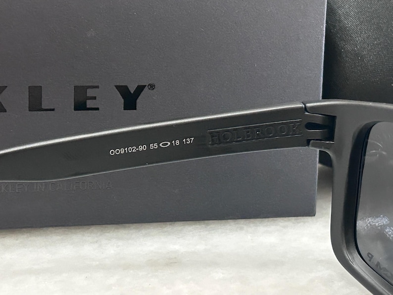 May include: Close-up of black Oakley Holbrook sunglasses. The frame features the model number OO9102-90 55 18 137. The word "HOLBROOK" is printed on the arm. The background is a black box with the Oakley logo.