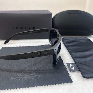 May include: Black Oakley sunglasses with a matching case, box, and cleaning cloth. The sunglasses have a sleek, modern design with dark lenses and a matte black frame. The Oakley logo is visible on the frame and accessories.