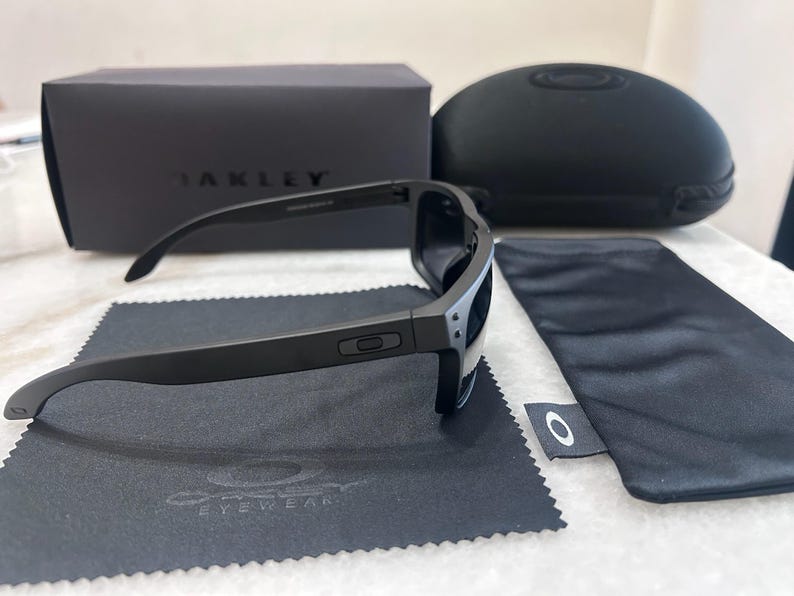 May include: Black Oakley sunglasses with a matching case, box, and cleaning cloth. The sunglasses have a sleek, modern design with dark lenses and a matte black frame. The Oakley logo is visible on the frame and accessories.