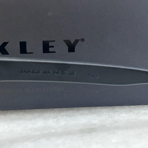 May include: Close-up of black sunglasses with the text "MADE IN USA" and the brand name "OAKLEY" visible. The sunglasses are set against a dark background. The text "DESIGNED BY OAKLEY IN CALIFORNIA" is also visible.