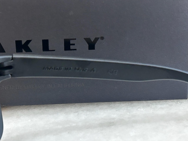 May include: Close-up of black sunglasses with the text "MADE IN USA" and the brand name "OAKLEY" visible. The sunglasses are set against a dark background. The text "DESIGNED BY OAKLEY IN CALIFORNIA" is also visible.
