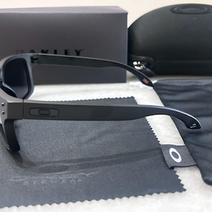 May include: Black Oakley sunglasses with a matching case, soft pouch, and cleaning cloth. The sunglasses have a rectangular frame and dark lenses. The Oakley logo is visible on the frame and accessories. The box is black with the Oakley logo.