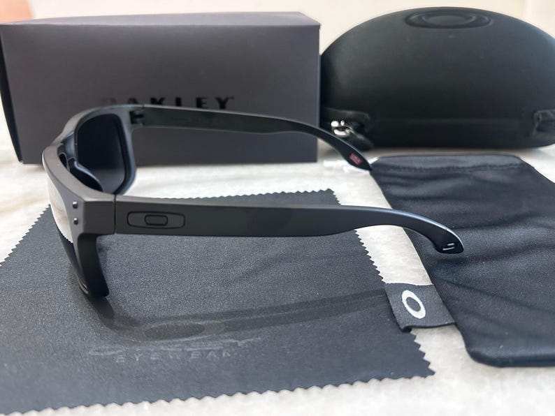 May include: Black Oakley sunglasses with a matching case, soft pouch, and cleaning cloth. The sunglasses have a rectangular frame and dark lenses. The Oakley logo is visible on the frame and accessories. The box is black with the Oakley logo.