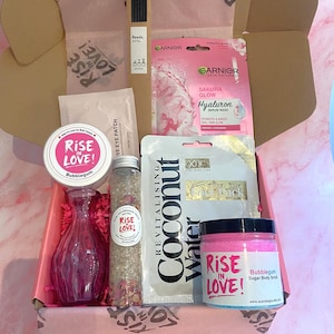 Op de afbeelding: Een roze geschenkdoos gevuld met schoonheidsproducten. Items zijn onder meer een roze glazen vaas, een bubblegum suiker bodyscrub, een gezichtsmasker, wierookstokjes en badzout. De doos is gevoerd met roze vloeipapier.