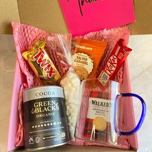 May include: A gift box containing a selection of treats: a Twix bar, Walker's salted caramel biscuits, a Kit Kat, a mug, cocoa, and marshmallows. A pink card with "Thanks" in gold lettering is also included.