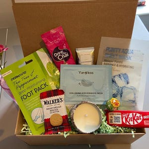 Op de afbeelding: Een cadeaupakket gevuld met zelfzorgartikelen. Inclusief een voetpakking, oogpatches, een kaars, een Kit Kat-reep, shortbread koekjes en een chai thee zakje. De doos is gevoerd met groen papier en heeft een takje roze bloemen.