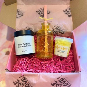 May include: A gift box with pink shredded paper, containing a jar of shea body butter, a decorative glass bottle with a straw, and a container of whipped soap. The box and tissue paper are printed with "Rise in Love!"