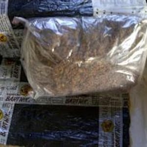 Pode incluir: Um saco plástico transparente cheio de grânulos castanhos está em cima de vários pacotes embrulhados. Os pacotes são pretos e presos com fita adesiva com a palavra "BARTNIK". O fundo é de cor amarelo claro.