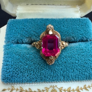 May include: A gold ring featuring a large, rectangular, faceted hot pink gemstone set in a decorative metal mounting with engraved floral details at the top and bottom, showcasing a vibrant colour contrast against the metallic gold band.