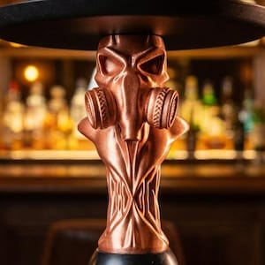 May include: A copper-coloured hookah with a gas mask design. The hookah has a black tray and a wooden mouthpiece. The background is a dimly lit bar with bottles of liquor on shelves.