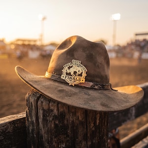 Peut inclure: Un chapeau de cowboy en feutre marron avec un emblème en métal doré sur la bande avant. L'emblème affiche un graphique de cheval et cavalier au galop avec le texte PRCA CHAMPION 2024 FINALS. Le chapeau a un large bord incurvé et une bande texturée.