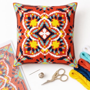 May include: A square throw pillow featuring a cross-stitch pattern with a central geometric motif in orange, yellow, turquoise, white, and dark blue. Beside it are a needle, a metal thimble, embroidery fabric, and several skeins of colored thread.