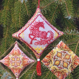 May include: Diamond-shaped Christmas ornament with a red horse design, beaded trim, and a red tassel. The ornament is displayed on a green pine tree branch. Two additional ornaments with geometric patterns are also visible.