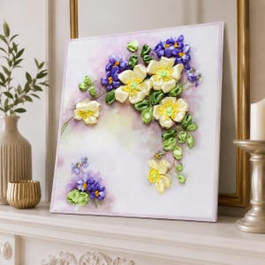 Può includere: Un'opera d'arte da parete quadrata con ricamo tridimensionale a nastro di fiori giallo pallido e piccoli fiori viola su uno sfondo in tessuto color viola chiaro e bianco in stile acquerello, con foglie e steli a nastro verde.