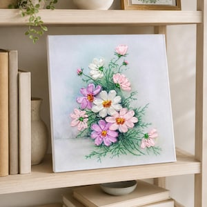 May include: A square canvas wall art piece featuring a textured ribbon embroidery design of cosmos flowers in shades of white, light pink, and lavender. The floral arrangement includes green embroidered stems and leaves on a pale, soft blue background.