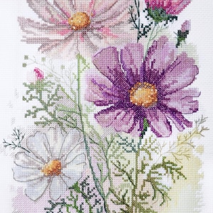 May include: Cross-stitch floral design featuring cosmos flowers in shades of pink, purple, and white. The flowers have yellow centers and are surrounded by green foliage, all on a white background.