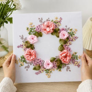May include: A square white canvas featuring a central wreath design created with pink and peach ribbon embroidery. The floral arrangement includes layered ribbon roses, small purple clusters, and green leaves, all stitched onto the fabric surface.