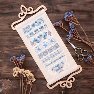 May include: A cross-stitch sampler with blue floral and geometric patterns, displayed on a wooden scroll. The embroidery features various shades of blue and beige, with a wooden hanger at the top and bottom. Dried flowers and scissors are also visible.