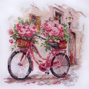 May include: Cross-stitch image of a pink bicycle with baskets overflowing with pink roses. The bicycle is parked on a cobblestone street in front of a building with a street lamp. The image has a soft, romantic feel.