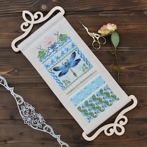 May include: A cross-stitch wall hanging with a blue dragonfly design. The embroidery features blue, green, and pink floral elements. The artwork is displayed on a beige fabric, held by a wooden scroll frame. Gold scissors and a dried rose are also visible.