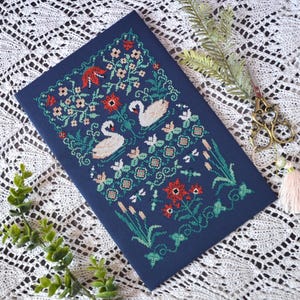 May include: A rectangular navy blue fabric panel featuring a detailed cross-stitch embroidery of two white swans swimming among red and pink flowers, green foliage, cattails, and dragonflies, accompanied by ornate metal scissors with a tassel.