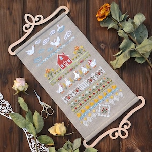 May include: A cross-stitch wall hanging depicting a rural scene with geese, a red house, and a white picket fence. The embroidery uses blue, yellow, green, and red threads, and is displayed on a wooden scroll frame.