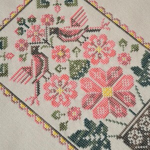 Puede incluir: Un bordado de punto de cruz sobre tela beige claro que presenta dos pájaros, varias flores rosas y hojas verdes. El diseño está encerrado dentro de un borde decorativo de pequeñas puntadas rosas y amarillas, creando un patrón rectangular.