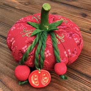 May include: A red tomato-shaped pincushion with green stem and leaves, and green braided accents. The pincushion has a floral pattern and two red felt ball accents, and a tomato slice pendant.