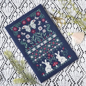 May include: A navy blue cross-stitch embroidery with a butterfly, flowers, and two rabbits. The design incorporates red, white, and green details. Gold scissors and a tassel are placed near the embroidery, which is on a white lace surface.