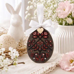 Op de afbeelding: Houten paasei-ornament met een kruissteekontwerp in rood en zwart. Het ei wordt opgehangen met een wit lint en een gouden hartvormige bedel. Een witte keramische konijn en bloemen staan op de achtergrond.