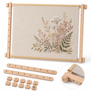 Op de afbeelding: Een rechthoekig houten borduurraam met een beige stoffen paneel voorzien van een gedetailleerd bloemenborduurwerk met witte, roze en groene wilde bloemen. De set bevat twee extra houten staven en tien kleine vierkante houten knoppen.