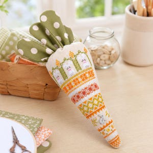 Può includere: Decorazione pasquale a forma di carota fatta a mano. La carota presenta un design a punto croce con coniglietti, motivi floreali e forme geometriche in arancione, bianco e verde. La carota ha foglie in tessuto verde a pois e un piccolo bottone a forma di coniglio.