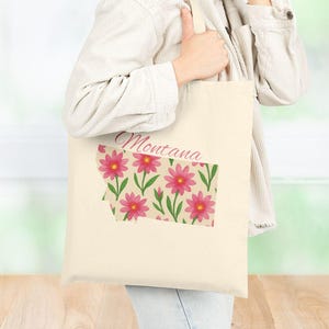 May include: Cream-colored tote bag featuring a floral design in the shape of Montana. The design includes pink flowers with green leaves and the word "Montana" in a cursive font. The bag is held by a person.