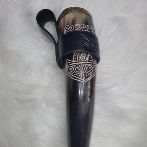 Peut inclure: Une corne à boire marron foncé avec des motifs gravés blancs et un support en cuir noir. La corne a un bord décoratif en laiton et est conçue pour être portée. La corne mesure environ 30 cm de long.