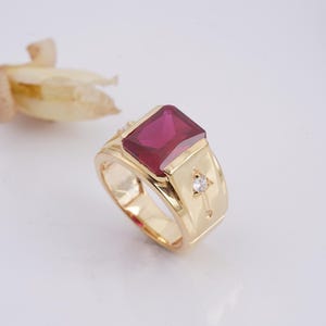 May include: A gold ring with a large, rectangular, ruby-colored gemstone. The ring has a thick band and is accented with small diamonds. The ring is set against a white background.