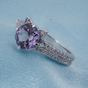 May include: A silver ring with a large, round, purple gemstone. The ring features a cat ear design on either side of the gemstone and a double band encrusted with small, clear stones. The ring is set against a light blue background.