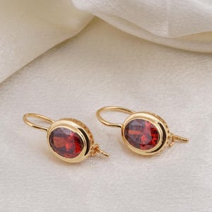 May include: A pair of gold earrings featuring oval, deep red gemstones. The earrings have a lever back closure and are set against a white, textured background. The gemstones are approximately 1.3 cm in length.