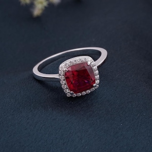 May include: A silver ring with a large, square-cut, deep red gemstone, encircled by a halo of smaller, clear stones. The ring's band is polished silver. The setting accentuates the rich colour of the central stone.