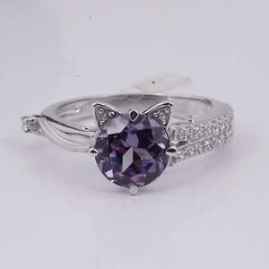 May include: A silver ring featuring a large, round, purple gemstone. The ring's design includes cat ears on the setting and a band with small, clear stones. The ring is set against a white background.