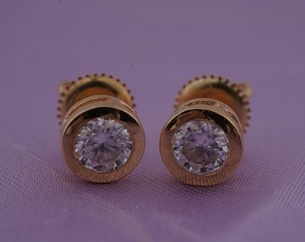 May include: A pair of gold-colored stud earrings with a round, clear gemstone set in a bezel. The earrings are displayed on a purple fabric surface.