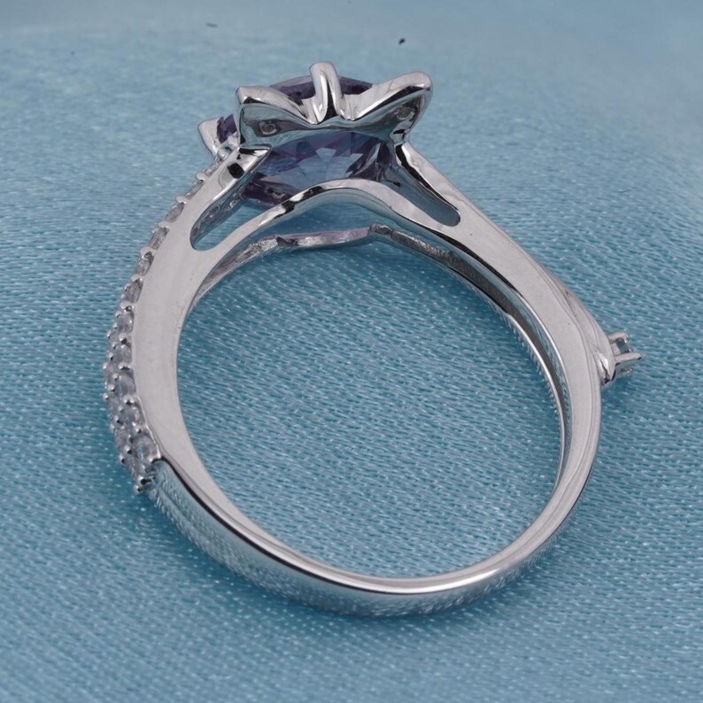 May include: A silver ring with a large, purple gemstone in a star-shaped setting. The band is adorned with small, clear stones along one side. The ring is set against a light blue background.
