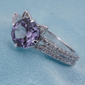 May include: A silver ring with a large, round, light purple gemstone. The ring features a cat ear design on either side of the gemstone and a double band encrusted with small, clear stones. The ring is set against a light blue background.