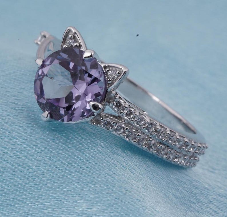 May include: A silver ring with a large, round, light purple gemstone. The ring features a cat ear design on either side of the gemstone and a double band encrusted with small, clear stones. The ring is set against a light blue background.