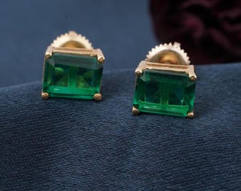 Pode incluir: Um par de brincos de botão dourados, cada um com uma pedra preciosa retangular verde esmeralda. Os brincos são colocados sobre um fundo de tecido azul escuro, mostrando sua cor e design vibrantes.