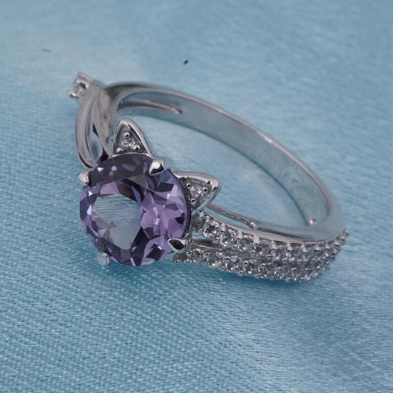 May include: A silver ring featuring a large, round, light purple gemstone. The ring has a cat-ear design on either side of the gemstone, with small, clear accent stones. The band is also adorned with small, clear stones.