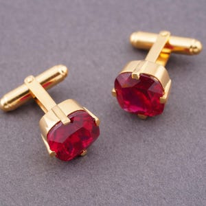 May include: A pair of gold-toned cufflinks with large, faceted, ruby-red gemstones. The cufflinks have a classic design with a rectangular gem setting and a T-bar closure. The background is a solid, neutral gray.