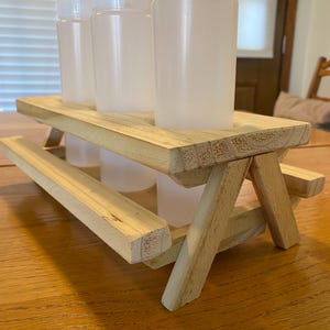 May include: A miniature wooden picnic table holds four clear plastic squeeze bottles with white caps. The table is made of unfinished wood and has a classic picnic table design. The bottles are empty and ready to be filled.