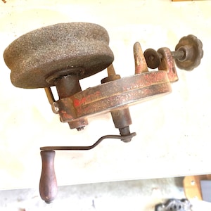 May include: An antique hand-cranked grinding wheel with a weathered, gray stone. The metal frame is painted red and shows signs of age. A wooden handle and a decorative knob are also visible.