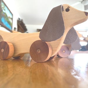 May include: A wooden dachshund toy with brown ears and wheels. The toy is made of light-colored wood with dark brown accents. The dachshund is positioned on a wooden surface.